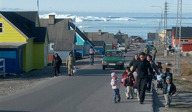 Daily life in a Greenlandic town with children walking and icebergs in the background, representing the culture and people affected by geopolitical tensions