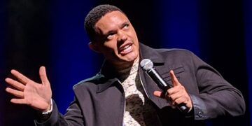 Comedian Trevor Noah performing on stage with a microphone, making an expressive face during a monologue